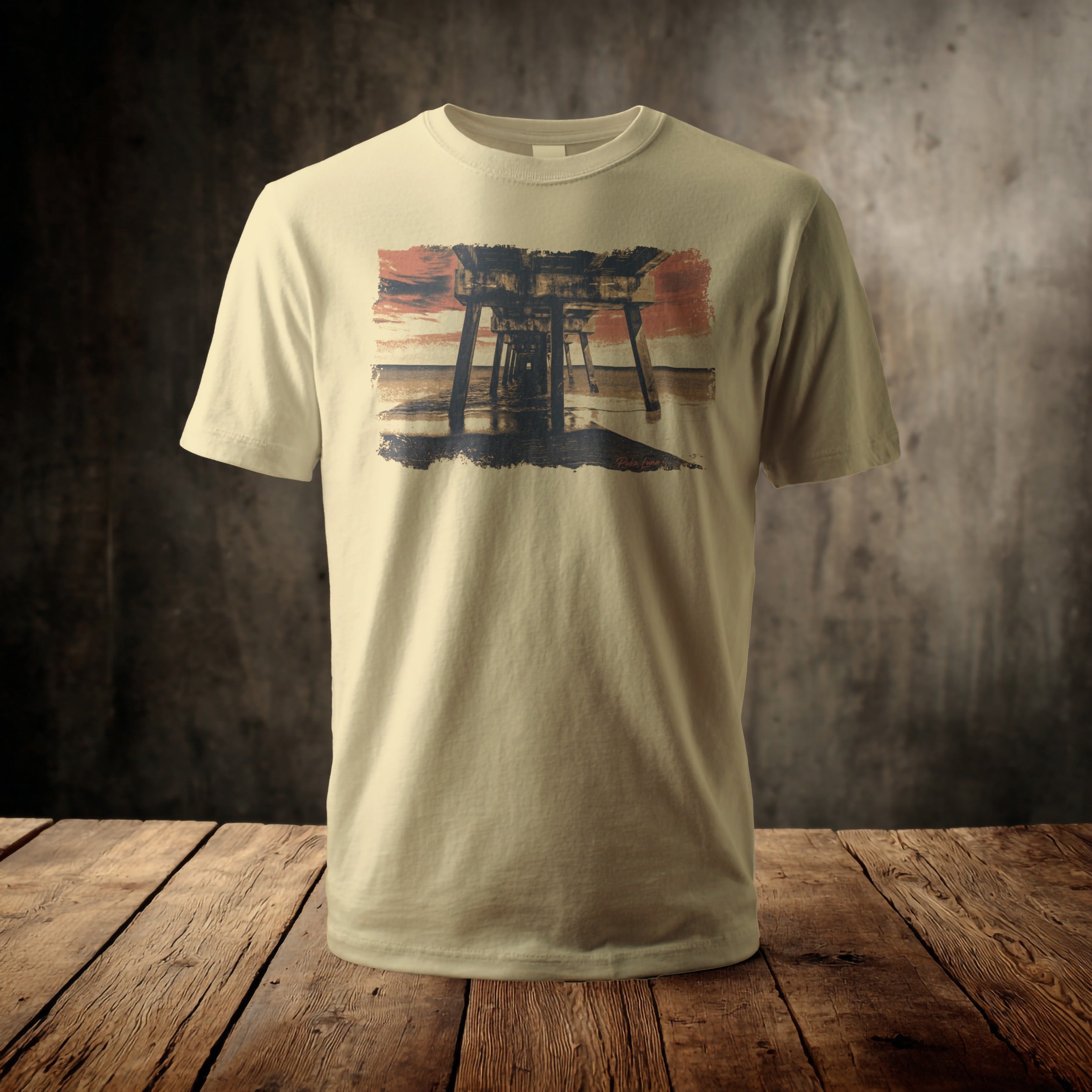 Light tan t-shirt with a sunset pier graphic on a wooden surface.