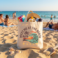 Canvas tote bag with "Meet Me at the Beach" graphic, beach scene, on sand.
