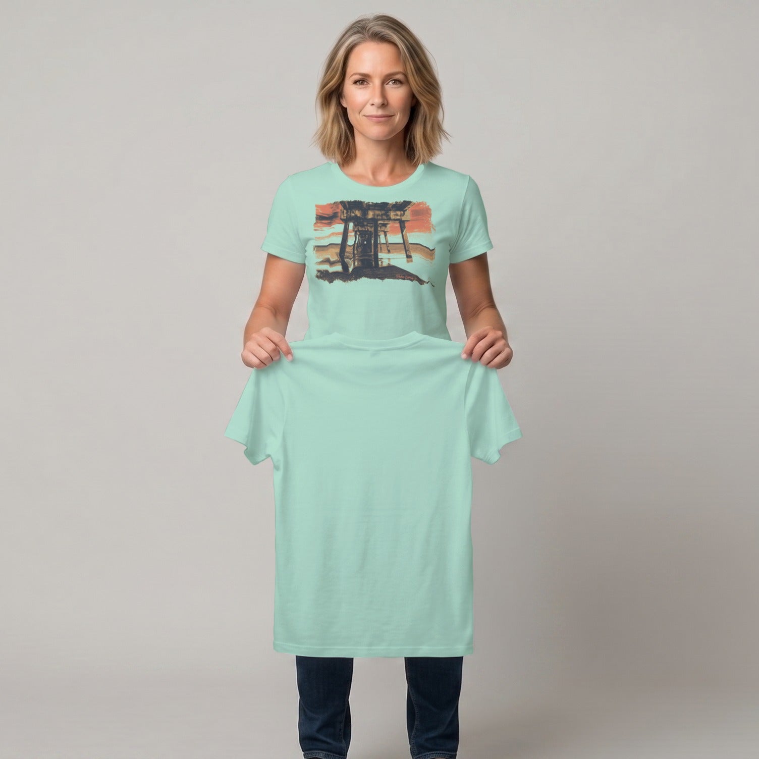 Woman holds up a mint green t-shirt with a sunset landscape graphic.