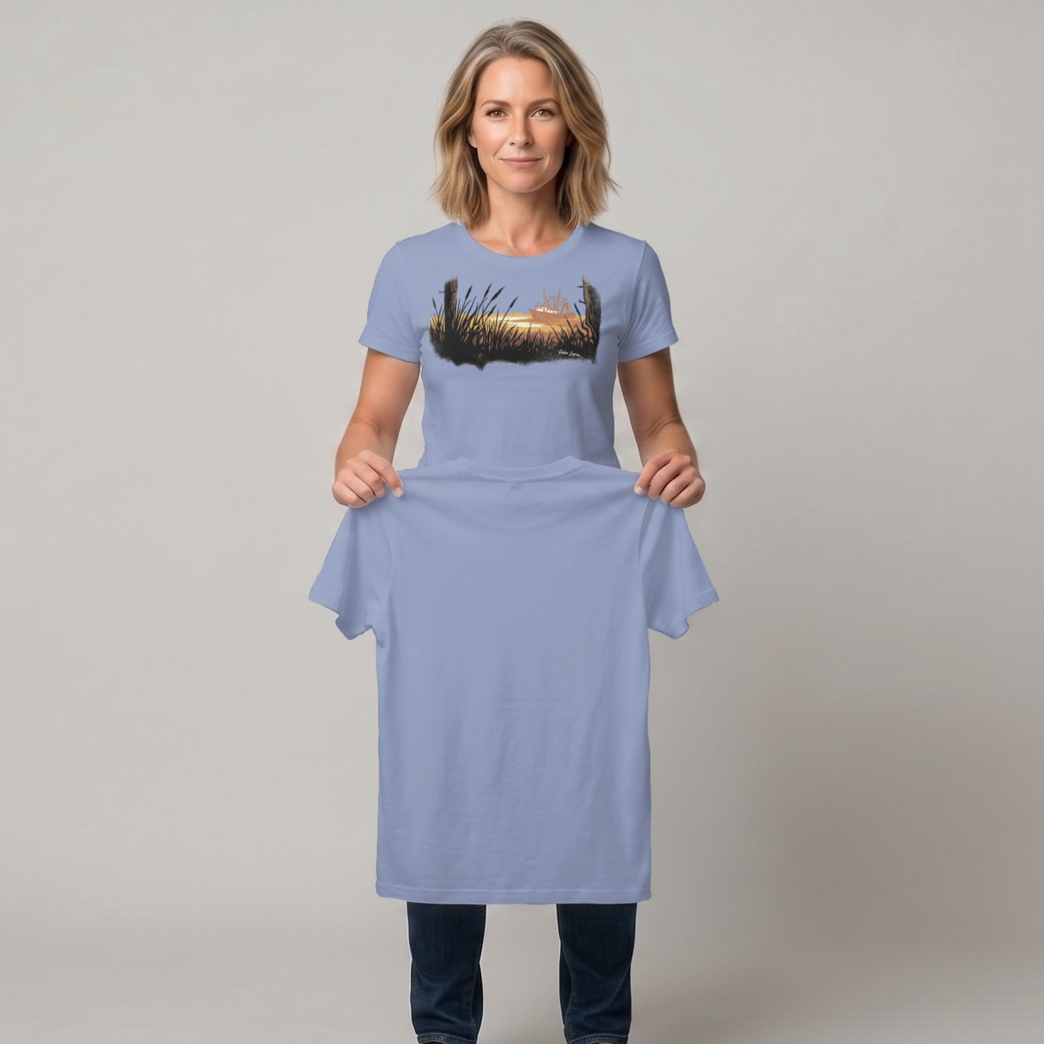 Woman holding up a light blue t-shirt with a sunset scene and grass graphic.