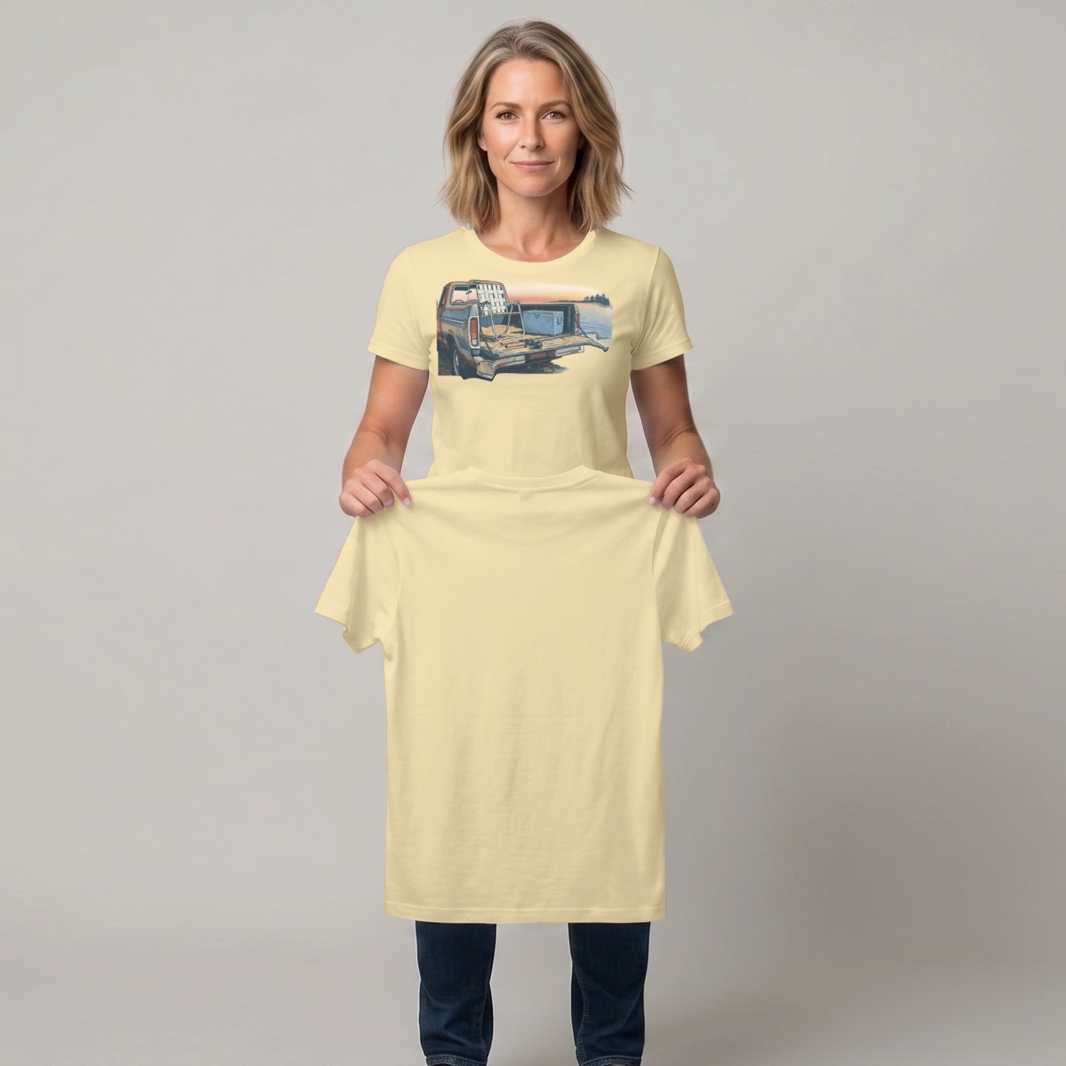 Woman holding a pale yellow t-shirt with a truck at sunset graphic.